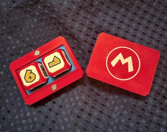 Op de afbeelding: Een rode rechthoekige plastic behuizing met twee witte dobbelstenen met gouden cijfers zes en één, naast een apart rood rechthoekig deksel met een wit cirkelvormig embleem met een hoofdletter M in het midden voor spelgebruik.