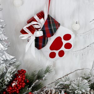 May include: A white stocking with a red paw print design and a plaid cuff. The stocking is filled with red and white wrapped gifts and is hanging from a red ribbon.