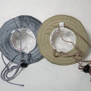Puede incluir: Dos sombreros de pescador, uno es azul claro y el otro es beige. Ambos sombreros tienen un forro blanco y un cord&oacute;n.