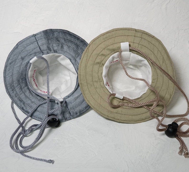 Puede incluir: Dos sombreros de pescador, uno es azul claro y el otro es beige. Ambos sombreros tienen un forro blanco y un cord&oacute;n.