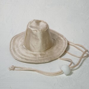 Puede incluir: Un sombrero de sol de color crema con un ala ancha y una correa para la barbilla. El sombrero est&aacute; hecho de una tela suave y sedosa.