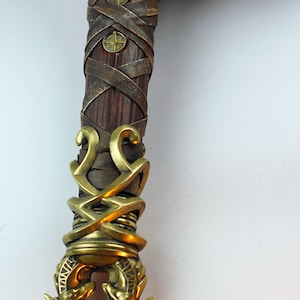 May include: Close-up of a sword hilt with intricate gold and brown leather details. The hilt is decorated with a woven leather wrap and a gold metal design with two animal heads at the bottom.