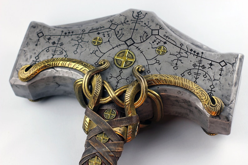 May include: A silver and gold hammer with a leather handle. The hammer head is decorated with intricate patterns and runes. The handle is wrapped in leather and has a gold metal band.