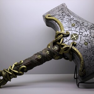 May include: A silver and gold hammer with a detailed design and intricate carvings. The hammer is on a black metal stand.