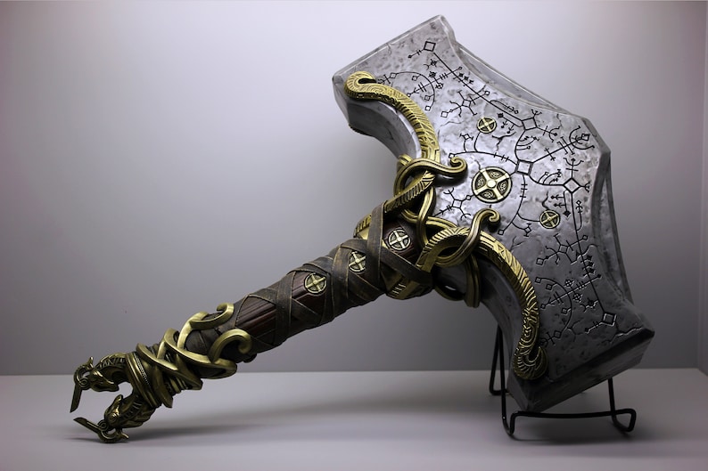 May include: A silver and gold hammer with a detailed design and intricate carvings. The hammer is on a black metal stand.