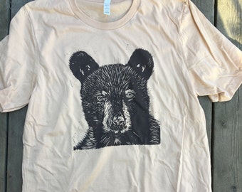Könnte beinhalten: Ein beiges T-Shirt mit einer Schwarz-Weiß-Illustration eines Bärenkopfes.