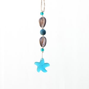 May include: A blue starfish charm hangs from a white string with brown and blue beads. The charm is a popular nautical theme for jewelry and home decor.
