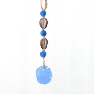 May include: A coastal-themed hanging ornament featuring a sand dollar, seashells, and blue glass beads strung on a natural twine. The sand dollar is a light blue color, and the seashells are brown.