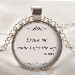 Op de afbeelding: Zilverkleurige hangerketting met een ronde bedel. De bedel toont de quote "Excuse me while I kiss the sky." in zwarte tekst, toegeschreven aan Jimi Hendrix, tegen een crèmekleurige achtergrond. De ketting heeft een fijne ketting.