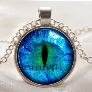 Op de afbeelding: Een zilveren ketting met een ronde hanger met een blauw en groen oogontwerp. De hanger heeft een zilveren rand en de tekst "Jewelly Wed" is zichtbaar aan de onderkant.