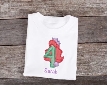 May include: White t-shirt with a green number 4 embroidered on it. The number is surrounded by red hair and a purple crown. There is a purple seashell below the number. The name "Sarah" is embroidered below the number.