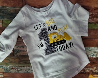 May include: White long-sleeved shirt with a playful design for a first birthday. The shirt features the text "Let's Dig and Play Today!" with a yellow and black construction vehicle graphic and the name "Declan". A large number 1 is also included.