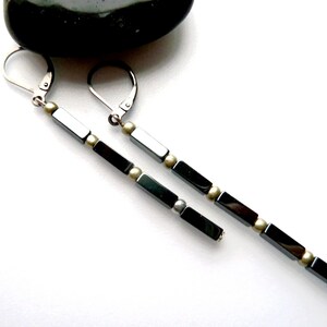 May include: A pair of long, dangling earrings with a linear design. The earrings feature alternating rectangular black beads and small, round, silver-coloured beads. The ear wires are silver-coloured. The earrings are displayed on a white background.