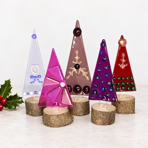 May include: Five triangular fused glass ornaments in light blue, magenta, mauve, purple, and red, each mounted on a small circular wooden log base. The glass pieces feature decorative glass beads, gold painted accents, and geometric patterns.