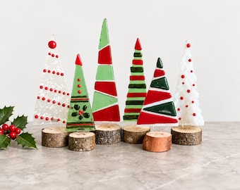 Peut inclure: Une collection de six ornements en verre triangulaires en forme d'arbres, chacun monté sur une petite base en bois circulaire. Les arbres présentent divers motifs en rouge, vert et blanc, incluant des rayures, des points et des motifs floraux.