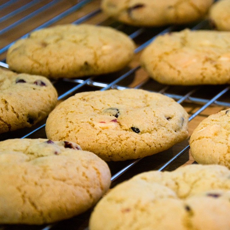 May include: Close-up of freshly baked cookies cooling on a metal rack. The cookies are golden brown with visible inclusions of dark and light-colored ingredients. The rack is made of silver-colored metal, and the background is a warm, wooden surface.