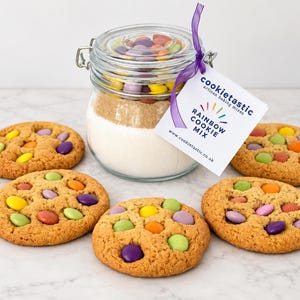 May include: A clear glass jar with a metal latch lid filled with layered dry cookie ingredients and colourful candy-coated chocolate pieces. A white tag reads cookietastic, RAINBOW COOKIE MIX, and a website. Four baked cookies with candy pieces sit nearby.