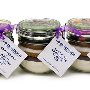 May include: Three glass jars filled with cookie mix ingredients, each with a different flavor: Cranberry & White Chocolate, Death by Chocolate, and Chocolate Orange. Each jar has a lid, a purple ribbon, and a tag with the flavor name.