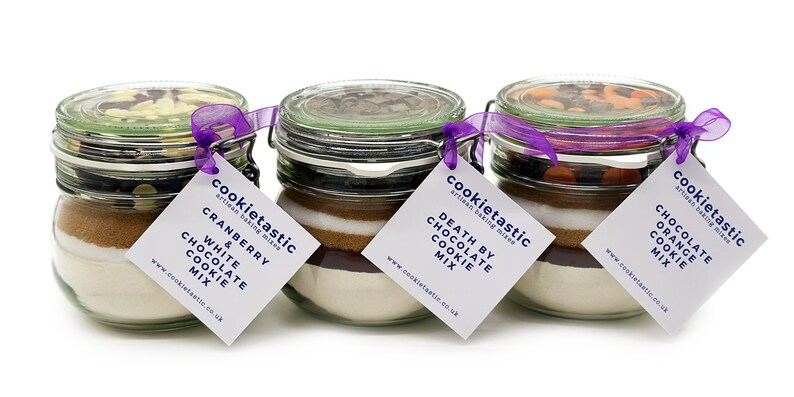 May include: Three glass jars filled with cookie mix ingredients, each with a different flavor: Cranberry & White Chocolate, Death by Chocolate, and Chocolate Orange. Each jar has a lid, a purple ribbon, and a tag with the flavor name.