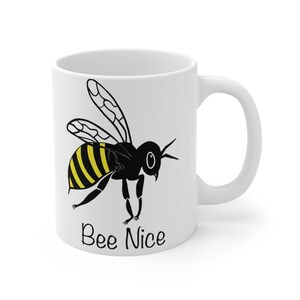 May include: White ceramic mug featuring a cartoon bee with yellow and black stripes and the words "Bee Nice" in black. The bee has detailed wings and is positioned on the side of the mug. The mug has a curved handle.