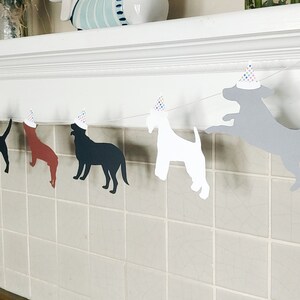 Op de afbeelding: Een slinger met silhouetten van honden in verschillende kleuren, elk met een feesthoedje. De slinger is gespannen over een wit oppervlak. Op de achtergrond zijn kleurrijke bloemen te zien. Perfect voor een feestje met een hondenthema.