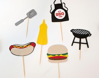 May include: A set of six barbecue-themed paper cupcake toppers on wooden picks, including a gray spatula, a black apron with the text IT'S BBQ TIME, a yellow mustard bottle, a hot dog, a hamburger, and a black charcoal grill on a stand.