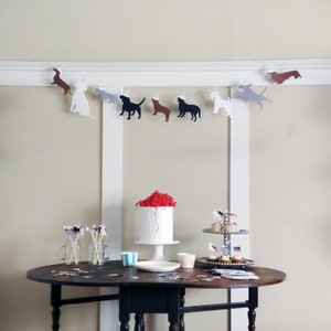 Op de afbeelding: Een feestelijke sc&egrave;ne met een hondenbanner, een witte taart met rode accenten en cupcakes op een etag&egrave;re. De banner toont verschillende honden silhouetten, sommige met feesthoedjes. De tafel is gedekt met feestartikelen.