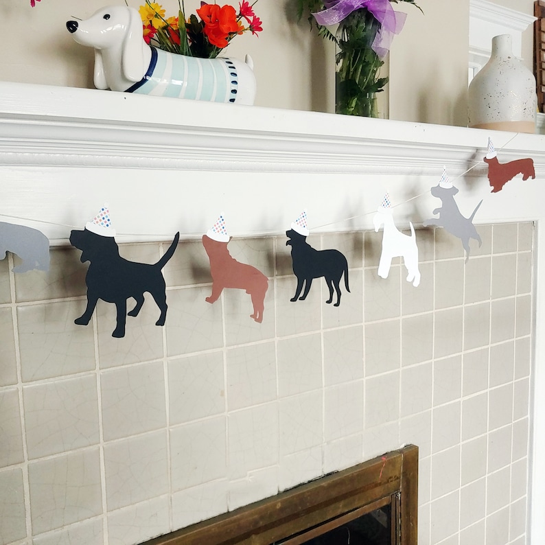 Op de afbeelding: Een feestelijke slinger met honden, met verschillende hondensilhouetten in zwart, bruin, wit en grijs, elk met een feesthoedje. De slinger is gespannen over een wit oppervlak, wat een feestelijke sfeer cre&euml;ert.