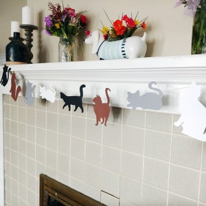 Op de afbeelding: Een slinger van katten silhouetten in verschillende kleuren, waaronder zwart, bruin, grijs en wit, hangt voor een betegelde muur. De slinger is gespannen over een witte schoorsteenmantel, versierd met bloemen en kaarsen.