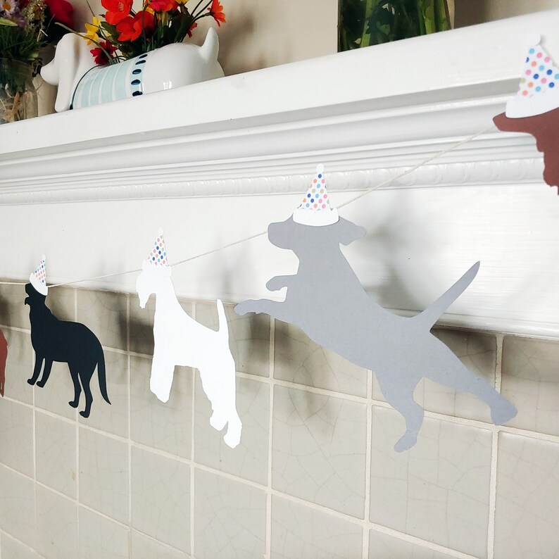 Op de afbeelding: Een feestbanner met hondenthema met silhouetten van honden in verschillende kleuren, elk met een feesthoedje met kleurrijke stippen. De banner is geregen aan een wit touwtje, perfect voor een verjaardag.