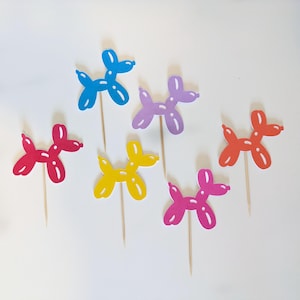 May include: Six colorful balloon animal cupcake toppers. The toppers are shaped like dogs and are made of paper. The colors are blue, purple, yellow, pink, red, and orange.