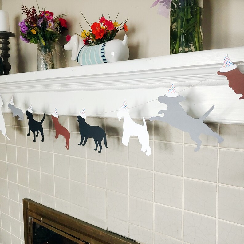 Op de afbeelding: Een slinger met silhouetten van honden in verschillende kleuren, elk met een feesthoedje. De slinger is gespannen over een wit oppervlak. Op de achtergrond zijn kleurrijke bloemen te zien. Perfect voor een feestje met een hondenthema.