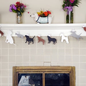 Op de afbeelding: Een feestelijke slinger met honden, met silhouetten van verschillende hondenrassen, elk met een feesthoedje. De honden zijn in verschillende kleuren, waaronder wit, grijs, bruin en zwart. De slinger hangt over een plank met bloemen in vazen.