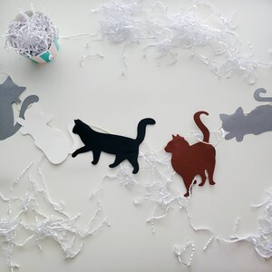 Op de afbeelding: Een slinger met katten als thema, met uitgesneden katten silhouetten in grijs, wit, zwart en bruin. De katten zijn geregen aan een witte draad, met wit papieren vulmateriaal eromheen. Een kleine, gestreepte container gevuld met papier staat in de linkerbovenhoek.
