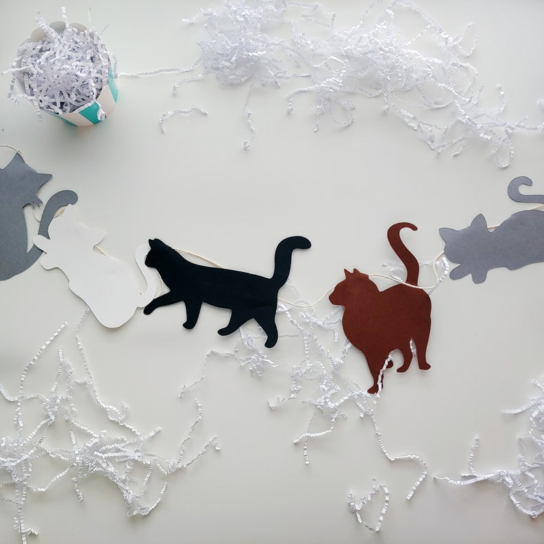 Op de afbeelding: Een slinger met katten als thema, met uitgesneden katten silhouetten in grijs, wit, zwart en bruin. De katten zijn geregen aan een witte draad, met wit papieren vulmateriaal eromheen. Een kleine, gestreepte container gevuld met papier staat in de linkerbovenhoek.