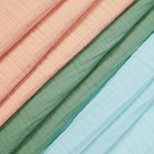 May include: Three layers of folded fabric in soft pastel tones. The top layer is a light peach, the middle layer is a muted sage green, and the bottom layer is a pale sky blue. Each section of the textile features a visible, crinkled, gauze-like texture.
