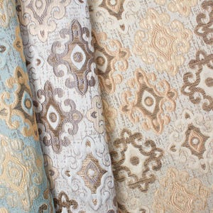 Pode incluir: Close-up de tecido estampado em tons de bege, castanho e azul claro. O tecido apresenta um design ornamental e repetitivo com uma aparência texturizada. O padrão inclui formas de diamante e florais, criando uma estética luxuosa.