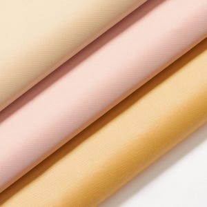 May include: Three layers of smooth, solid-colored fabric are folded diagonally across the frame. The top layer is a light beige, the middle layer is a soft pastel pink, and the bottom layer is a warm, golden yellow hue with a subtle sheen.