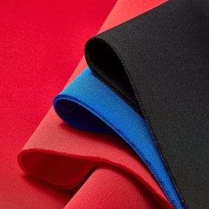 May include: A close-up of layered fabric in red, blue, and black. The red fabric is smooth, while the blue and black fabrics have a textured appearance. The blue fabric has a blue stitched edge.