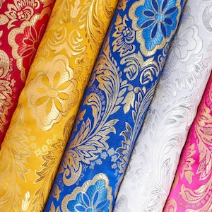 May include: Close-up of several rolls of fabric in various colors, including red, yellow, blue, white, and pink. Each roll features a gold floral damask pattern. The fabric appears to be a luxurious material, possibly for upholstery or drapery.