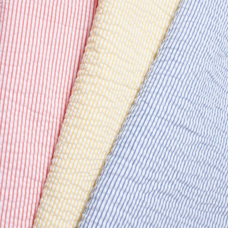 May include: A close-up of three sections of seersucker fabric in red and white stripes, yellow, and blue and white stripes. The fabric has a textured, puckered appearance, with vertical stripes. The colours are bright and vibrant, suitable for clothing or home decor.