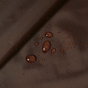 Può includere: Primo piano di tessuto marrone con gocce d'acqua. Il tessuto ha una trama strutturata e un colore marrone scuro. Diverse gocce d'acqua sono sparse sulla superficie, riflettendo la luce. L'immagine evidenzia le proprietà idrorepellenti del tessuto.