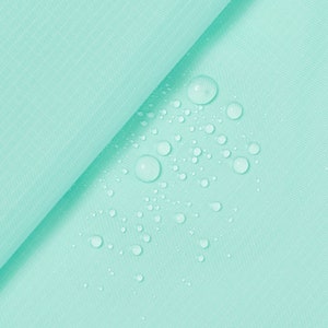 May include: A close-up of a light turquoise fabric with a grid pattern. Water droplets are scattered across the surface, creating a textured effect. The fabric is folded, revealing its thickness and texture.