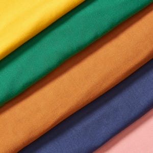 Pode incluir: Uma variedade de amostras de tecido coloridas em tons de amarelo, verde, laranja, azul marinho e rosa. O tecido está disposto diagonalmente, mostrando as diferentes cores e texturas do material.