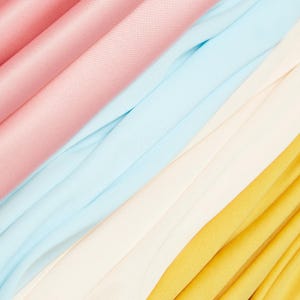 Op de afbeelding: Een abstracte afbeelding van gevouwen stof in pastelkleuren. De kleuren zijn roze, lichtblauw, crème en geel. De stof is diagonaal over het frame gerangschikt, wat een gevoel van beweging en textuur creëert.