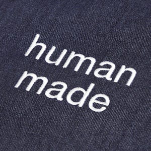 May include: Close-up of a dark blue denim fabric with the embroidered text "human made" in white. The text is in a sans-serif font and is centered on the fabric.