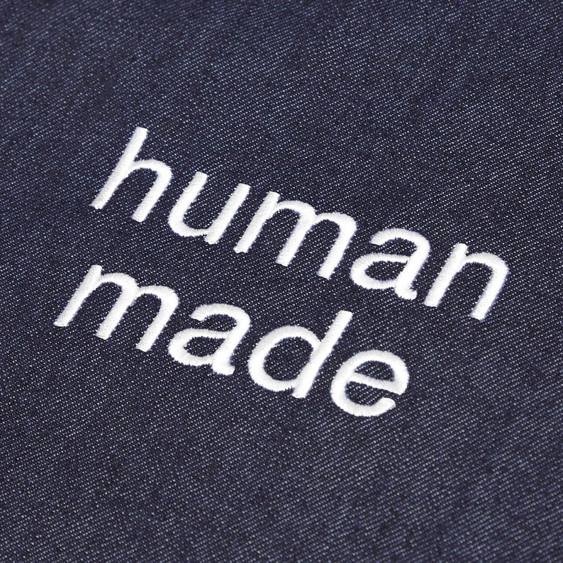 May include: Close-up of a dark blue denim fabric with the embroidered text "human made" in white. The text is in a sans-serif font and is centered on the fabric.