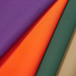 Op de afbeelding: Close-up van vier stoffen stalen in levendige kleuren. De stalen zijn paars, oranje, groen en beige. De stof heeft een gladde textuur en is gevouwen om de verschillende kleuren en texturen te laten zien. Ideaal voor naai- en knutselprojecten.