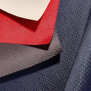 May include: A collection of four fabric swatches featuring a consistent hexagonal honeycomb texture. The layers are arranged in overlapping sheets of cream, bright red, dark brown, and deep navy blue, showcasing the uniform geometric pattern.