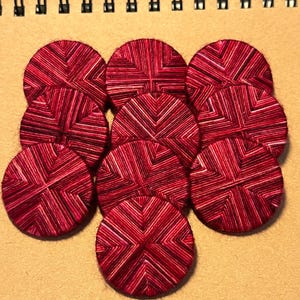 May include: Nine circular coasters in shades of red and burgundy, featuring a geometric pattern. The coasters are crafted from woven threads, providing a textured feel. They are arranged on a tan paper surface.